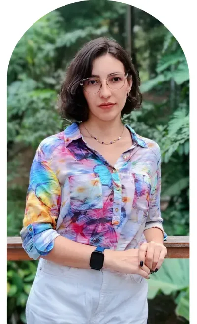 Dra. Letícia Aguiar em área externa, em pé, com fundo verde com folhagens, na seção sobre mim