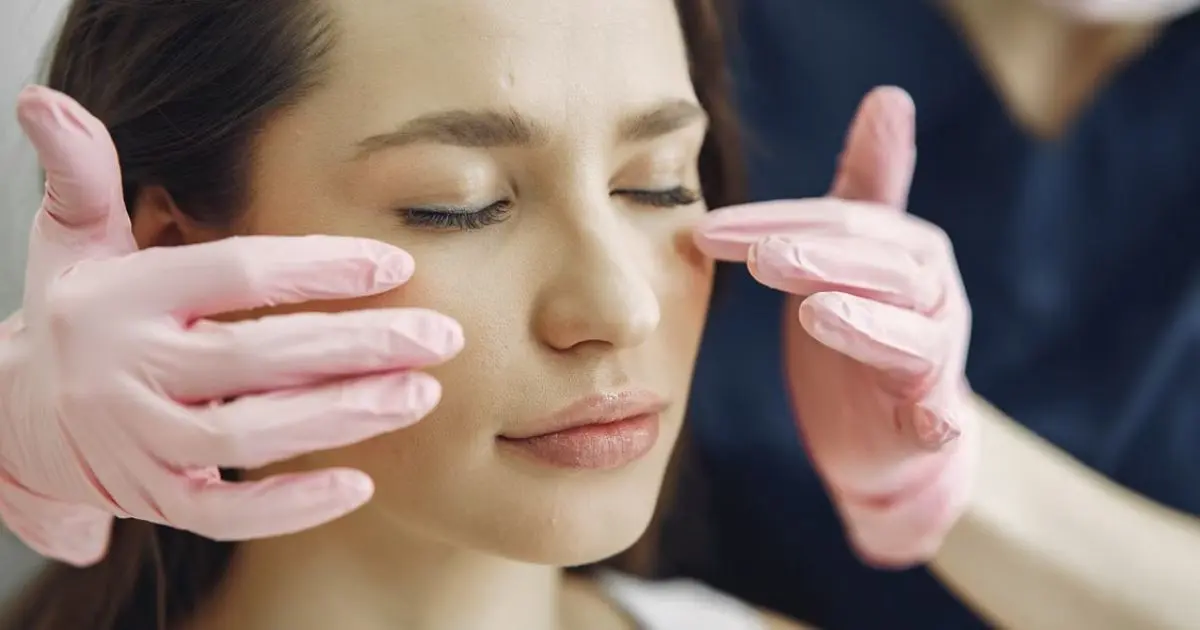 Mulher com os olhos fechados durante tratamento facial com as mãos de uma profissional posicionadas sobre o rosto