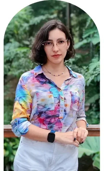 Dra. Letícia Aguiar em área externa, em pé, com fundo verde com folhagens, na seção sobre mim